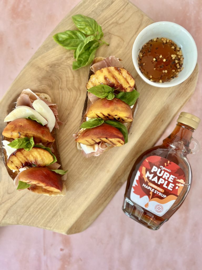 Grilled maple peaches on toasted sourdough bread with mozzarella, prosciutto, and basil, with a maple syrup bottle to the side and a bowl of maple syrup with chilli flakes