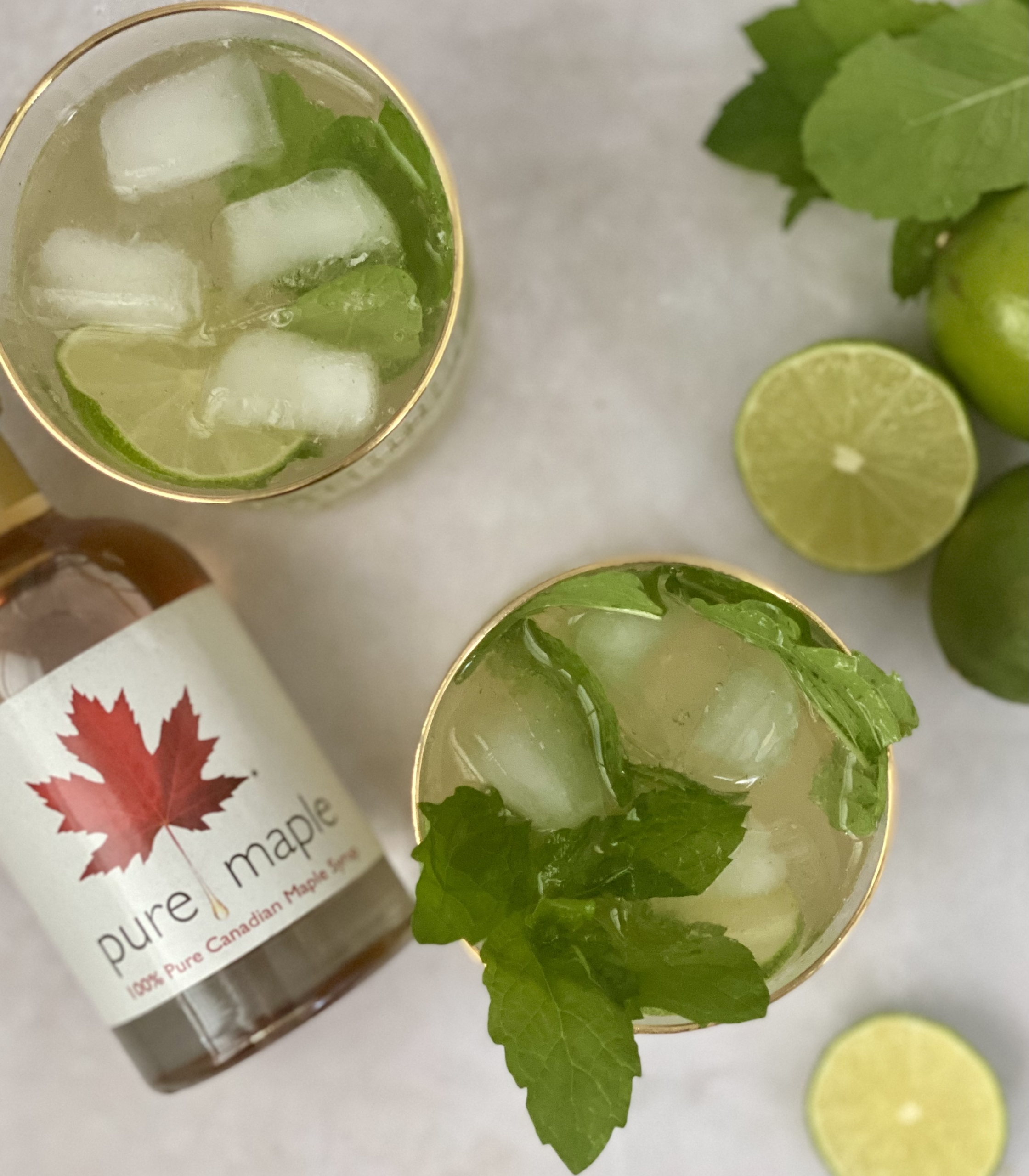 2 glasses of Mojitos with maple syrup bottle, limes and mint leaves