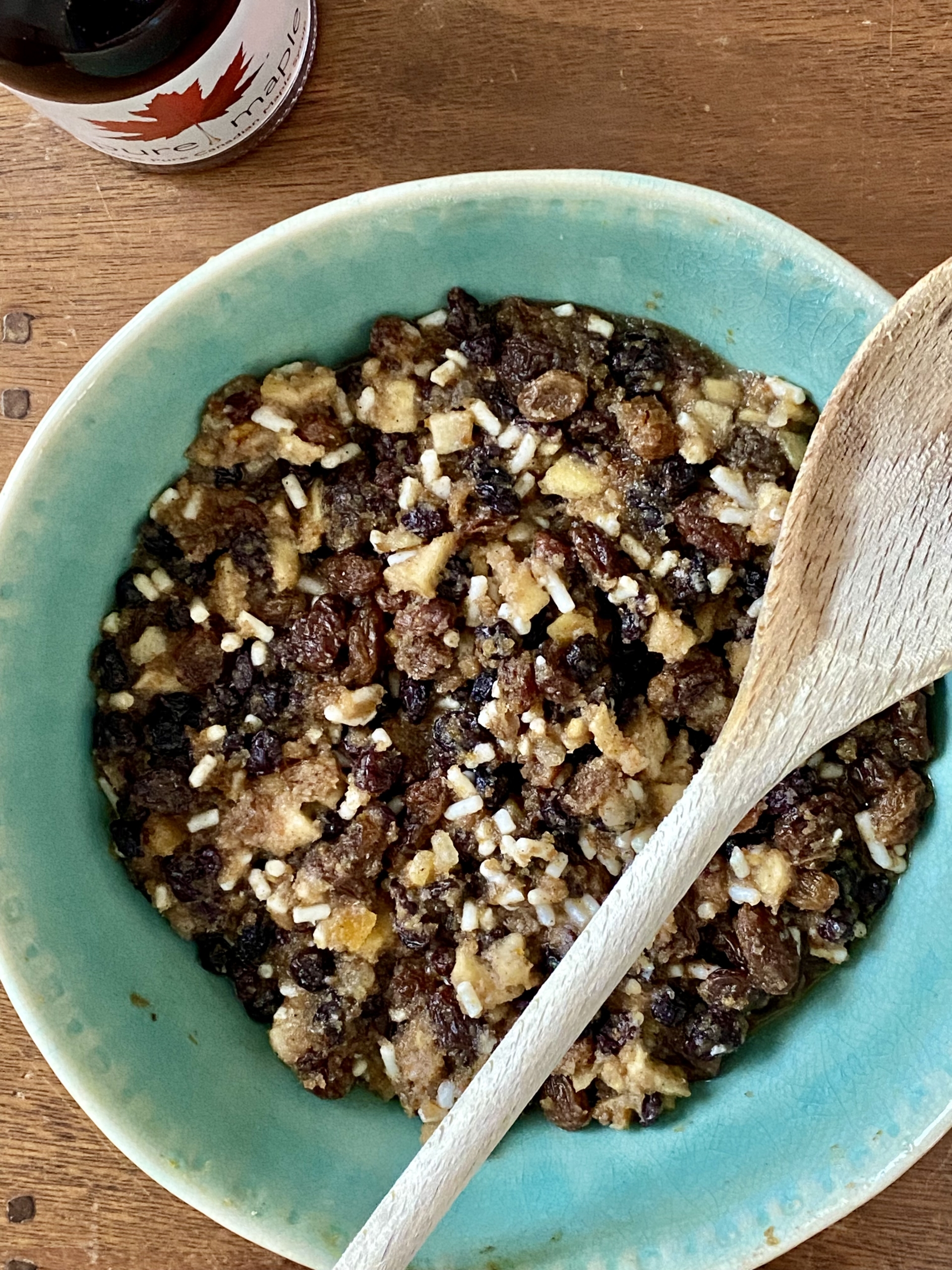 Christmas pudding in a large mixing bowl with a wooden spoon on the side and a bottle of maple syrup at the top